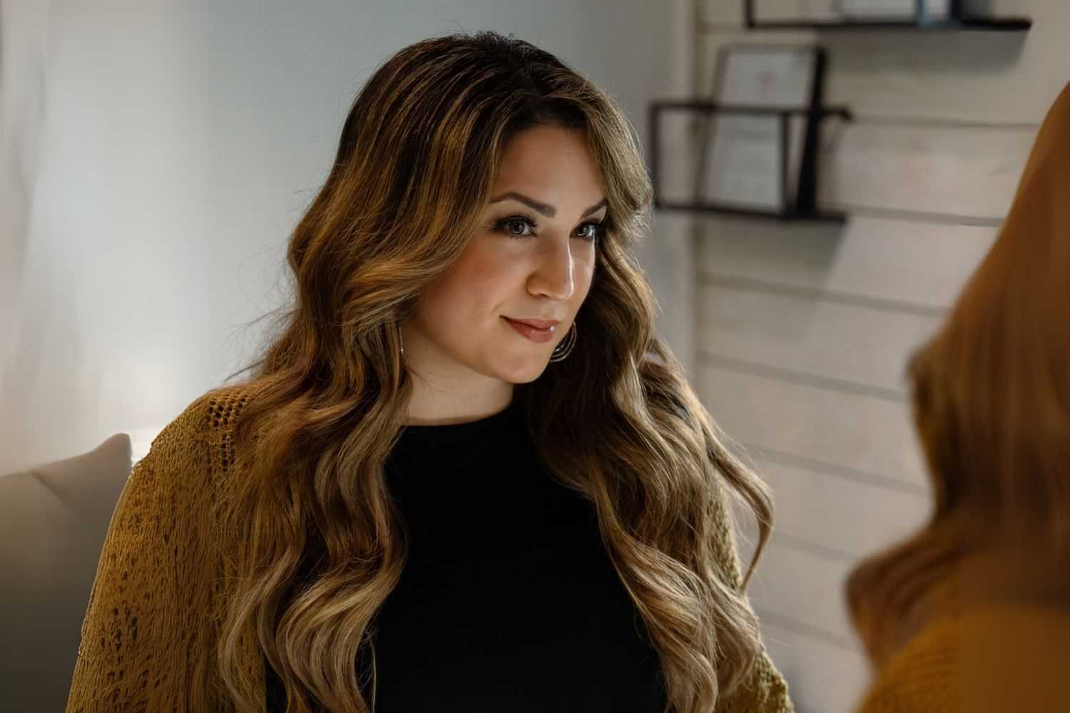 Woman with long hair smiling in a warmly lit room, gazing at a mirror.
