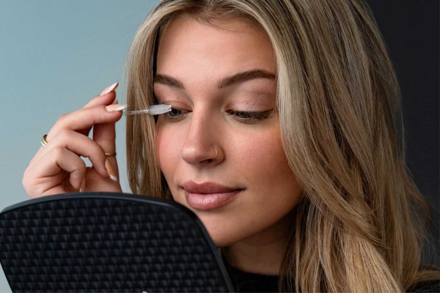 Woman applying mascara while holding a mirror.