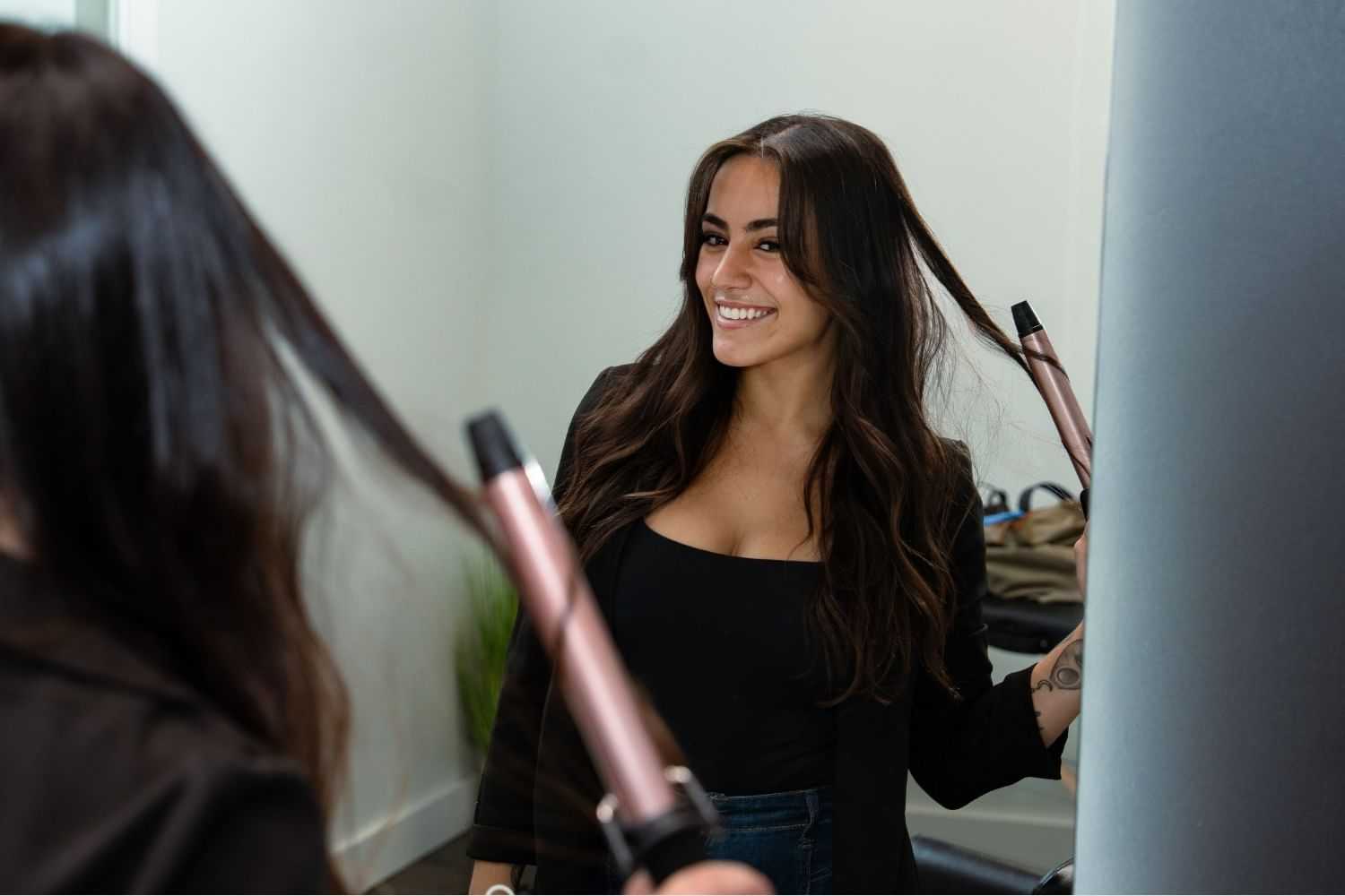 Woman curling her hair while smiling in front of a mirror.